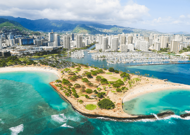 An aerial view of Downtown Honolulu, Hawaii, against a mountainous backdrop and coastal beaches and a marina in the foreground