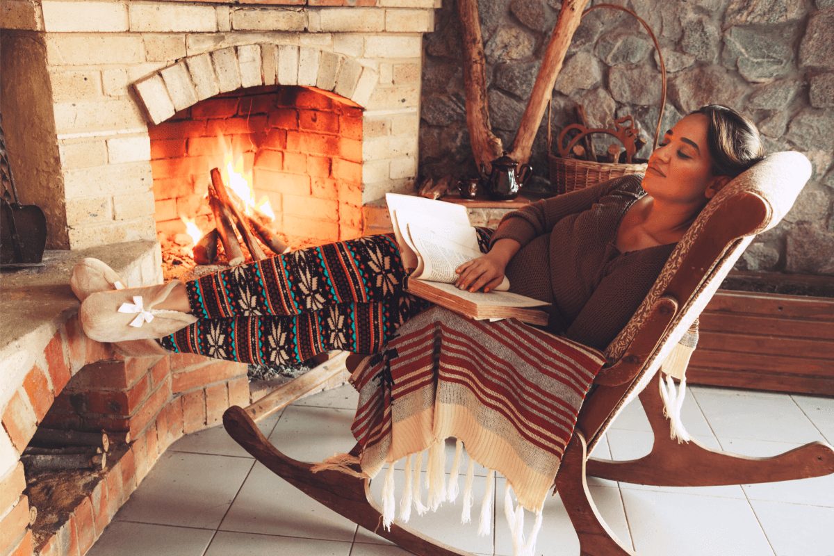 A woman relaxes by the fireplace in her pajamas while sitting in a rocking chair and reading a book